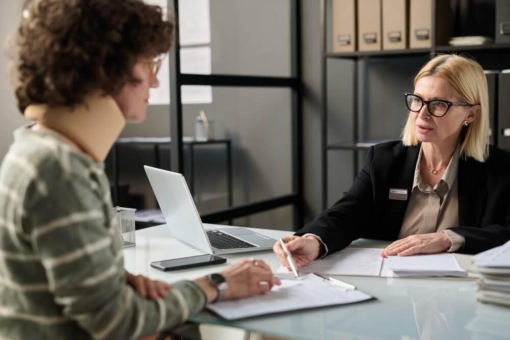 Portrait of female insurance broker consulting injured young woman in office after accident and pointing at legal forms
