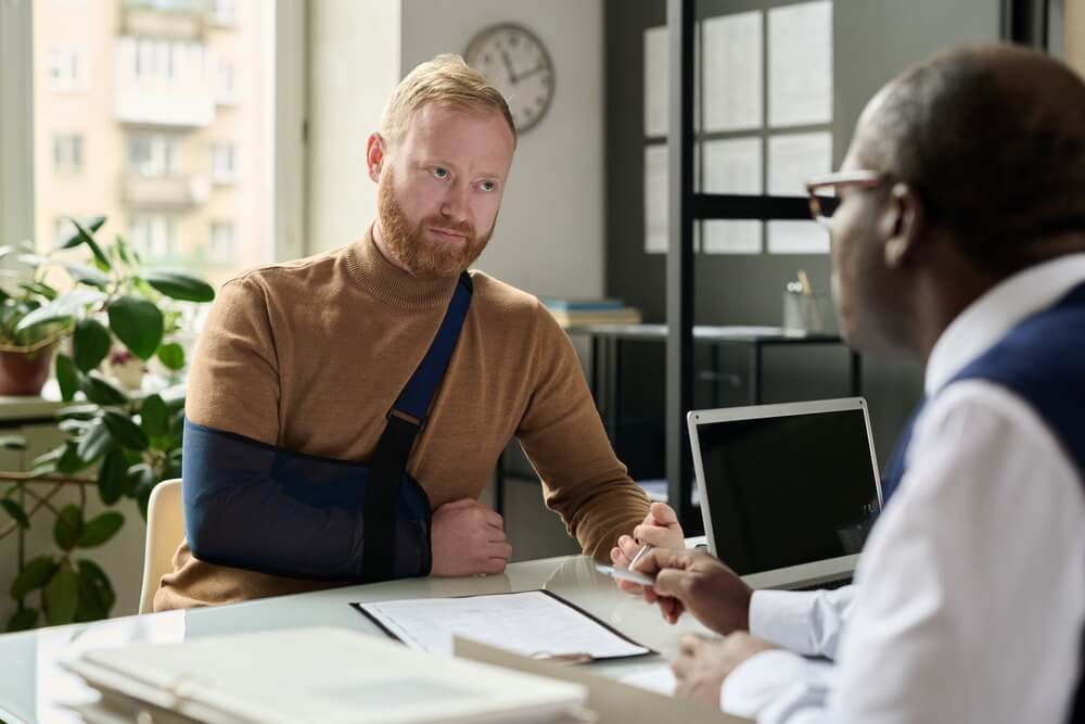 Portrait of man with hand in sling talking to insurance broker after workplace injury or accident
