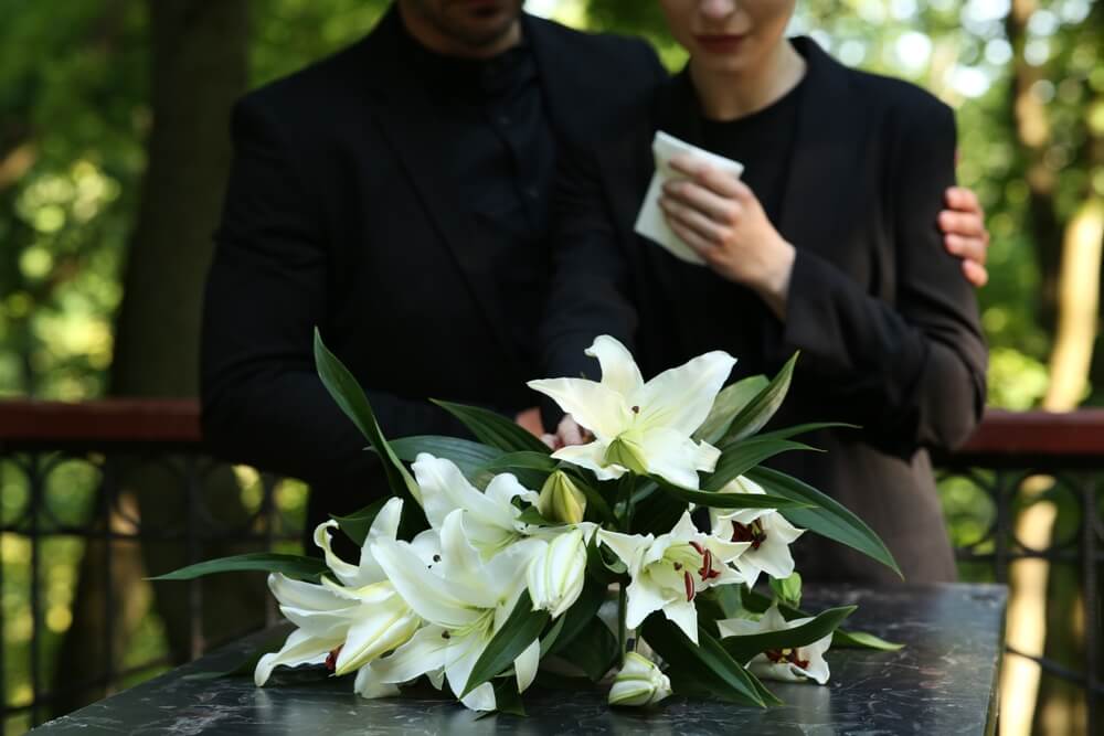 Couple near granite tombstone with white lilies at cemetery outdoors