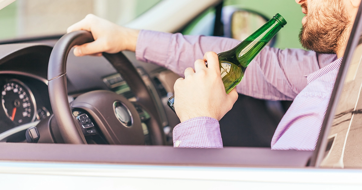 Man in a purple shirt holds a beer bottle with one hand while driving a car. The image conveys a sense of recklessness and danger.