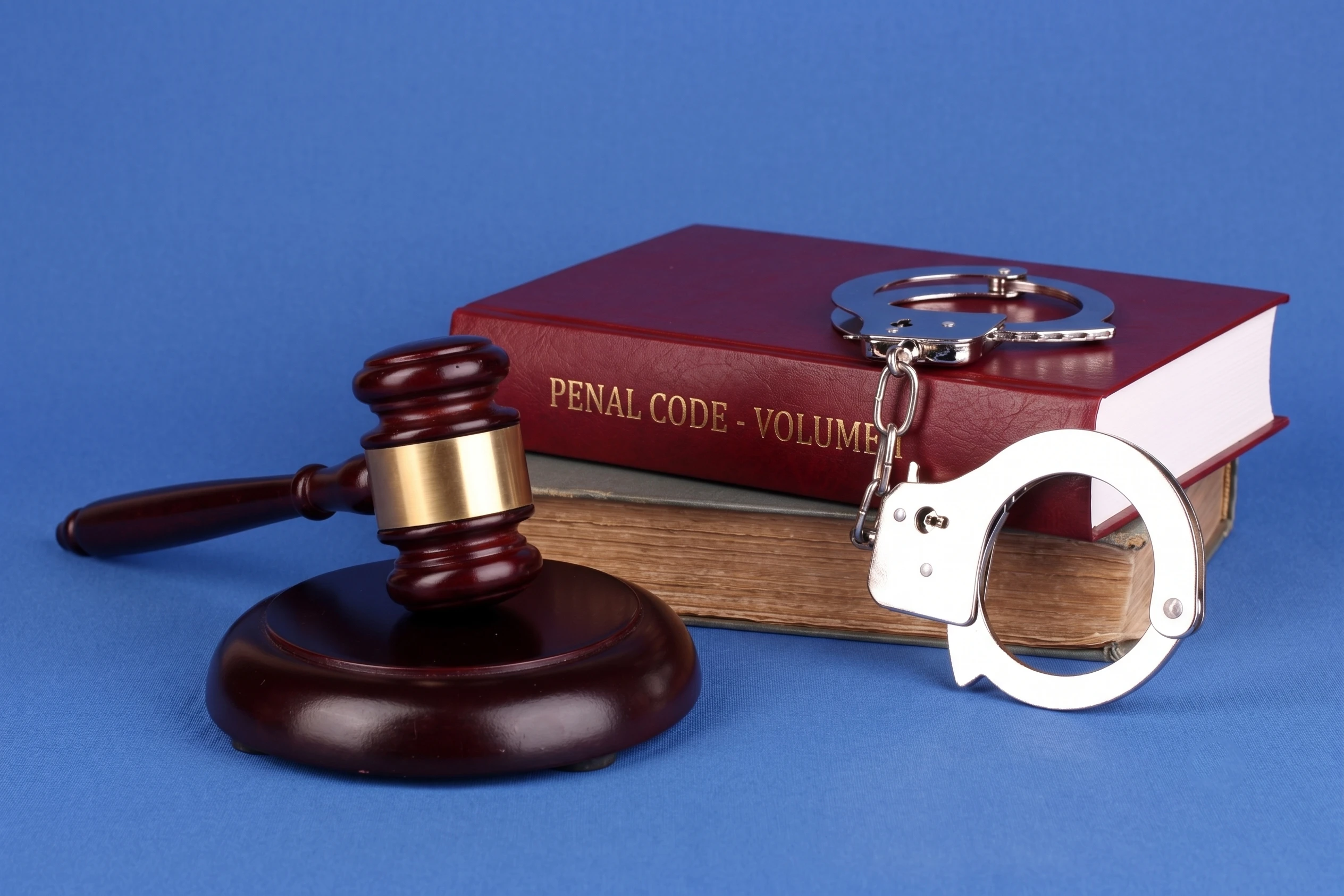 A wooden gavel, silver handcuffs, and a red "Penal Code" law book against a plain blue background.