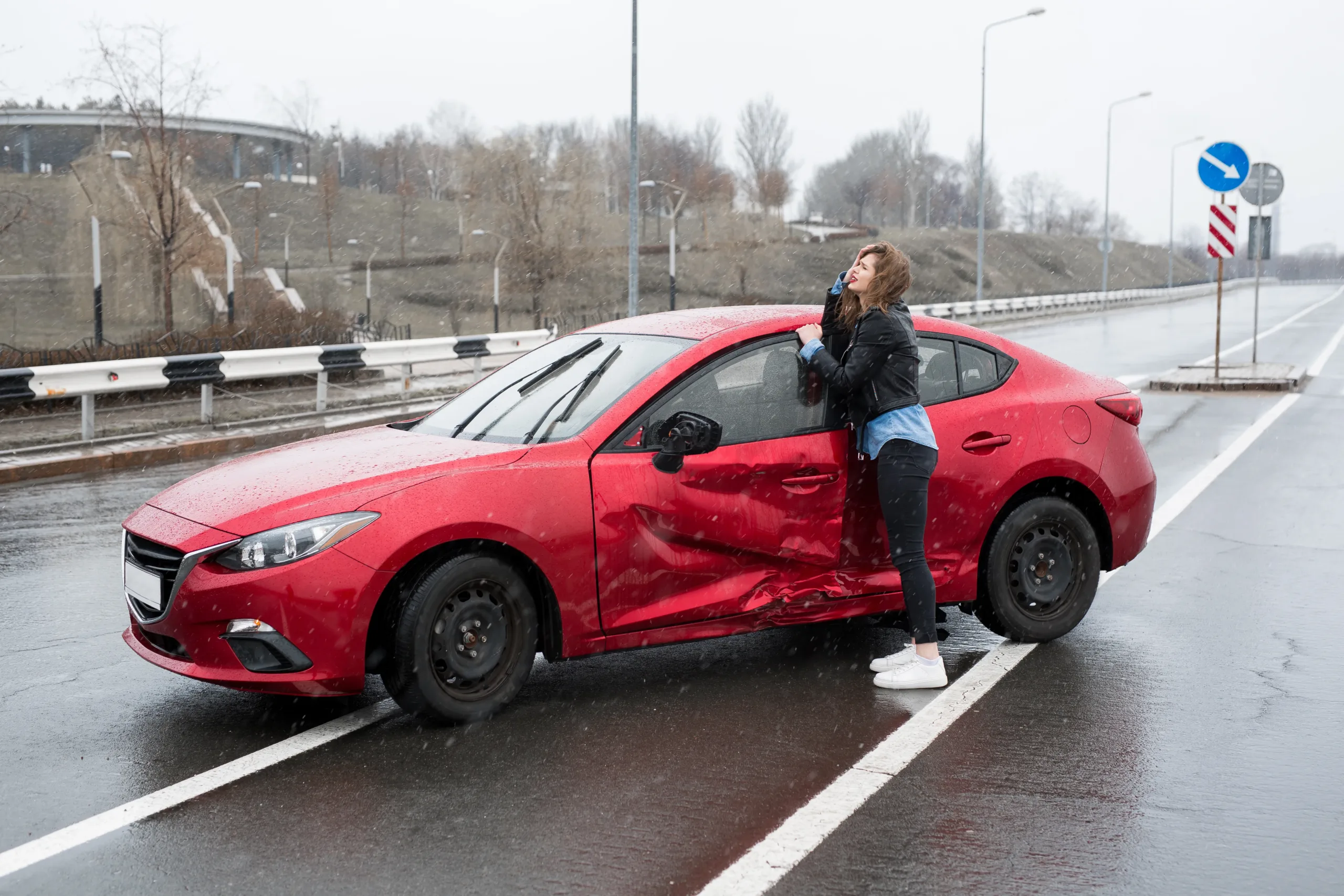 A distressed woman holding her head while standing beside a damaged red car on a wet road